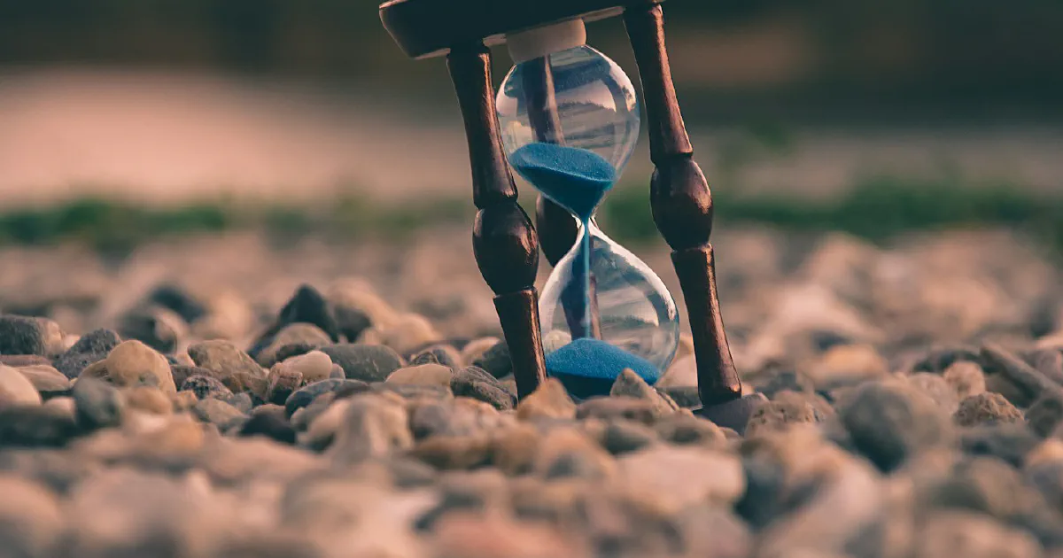 A small wooden hourglass with blue sand standing at a tilt on pebbles