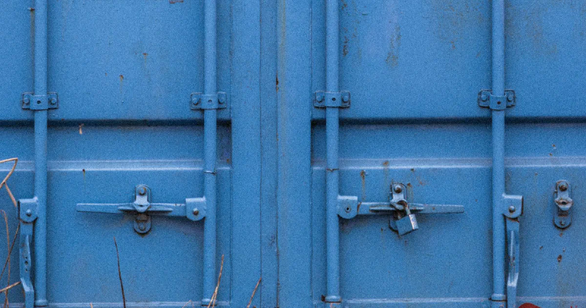 Close-up of the double doors of a weathered blue shipping container, with a small padlock hanging from the latch on the right door
